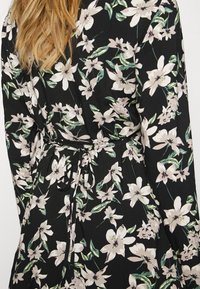 Blomklänning med svart bakgrund, prydd med stora blekrosa blommor och gröna blad. Designad med insvängd midja och knytdetalj.