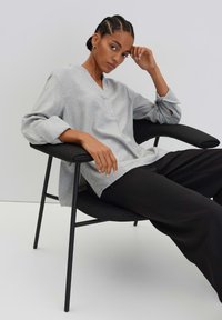 Tunique grise avec un décolleté en V et des manches retroussées, associée à un pantalon noir à jambe large, assise sur une chaise moderne noire. Design simple et décontracté.