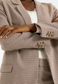 Blazer à motif pied-de-poule brun et crème avec revers crantés, trois boutons sur les poignets et une poche simple à l'avant.