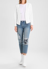 Bílý blazer, světlé tričko, modré džíny s nášivkami a výšivkou, a bílé kotníkové boty. Jednoduchý, moderní design s význačnými detaily.