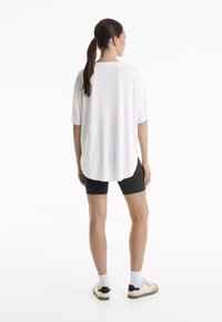 T-shirt blanche oversize avec un ourlet arrondi, associé à un short de vélo noir. Le mannequin porte des chaussettes blanches et des chaussures décontractées. Fond uni.