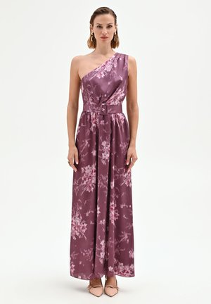 Femme portant une robe maxi fleurie violette sans manches, à une épaule, avec une ceinture assortie et des talons pointus couleur nude, debout sur un fond blanc.