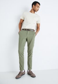 Camiseta color beige claro con un bolsillo en el pecho, pantalones verde oliva y zapatillas marrones con suelas oscuras; presenta un cinturón marrón y un diseño ajustado.