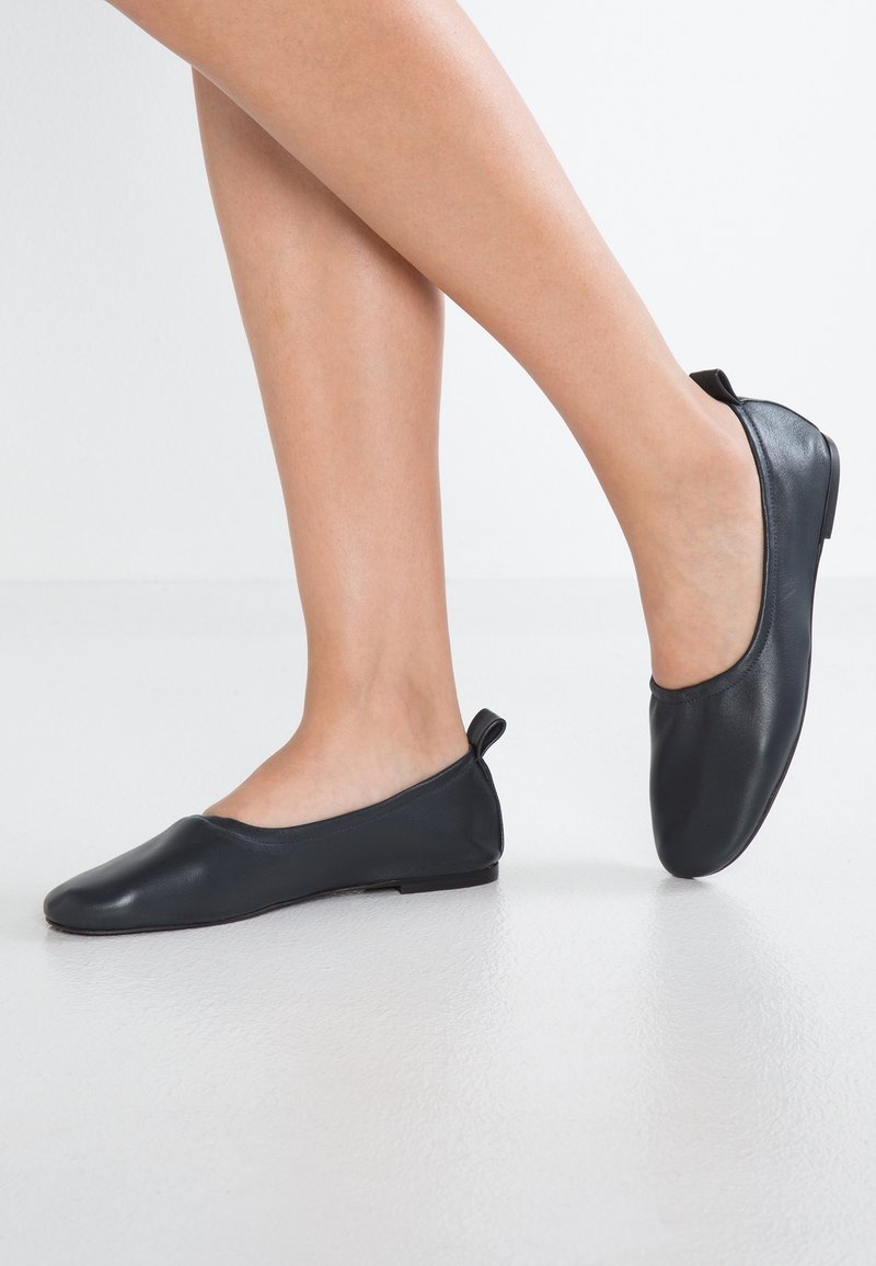 Ballerines en cuir noir à texture lisse et bout arrondi, avec une petite bride à l'arrière et un design minimaliste sur fond blanc.