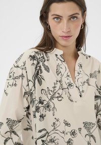 Cremefarvet bluse med et sort blomstermønster, der har en V-udskæring og afslappet pasform, lavet af letvægtsstof med en glat tekstur.