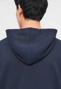 Άνδρας με κοντά μαύρα μαλλιά, φοράει σκούρο μπλε κουκούλα, ο οποίος φαίνεται από πίσω απέναντι σε ένα απλό φωτεινό φόντο.