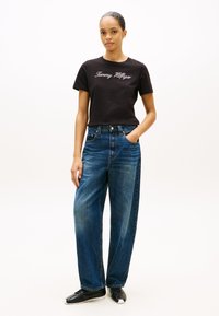 Musta puuvillainen T-paita, jossa "Tommy Hilfiger" kirjailtu valkoisella, yhdistetty löysiin, haalistuneen sinisiin leveälahkeisiin farkkuihin. Mustat tennarit jalassa.