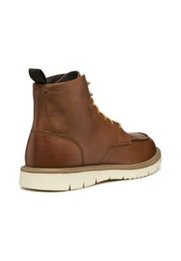 Bota de cuero marrón hasta el tobillo con cordones amarillos, un cuello acolchado y una suela de goma color crema. Incluye una lengüeta en el talón.