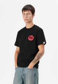 Tricou scurt cu mâneci negre, din bumbac, având un logo rotund roșu cu numărul "981" și textul "REPLAY". Material ușor texturat.