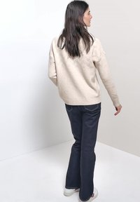 Frau mit langen dunklen Haaren, die einen cremefarbenen Strickpullover, dunkel ausgestellte Jeans und weiße Sneaker trägt und mit dem Rücken zu einem einfarbigen Hintergrund steht.