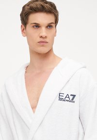 Accappatoio bianco in spugna con cappuccio, caratterizzato dal logo ricamato blu "EA7 EMPORIO ARMANI" sul petto, con una texture morbida e pelosa.
