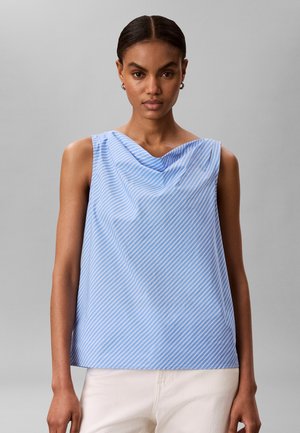 Vrouw draagt een mouwloze blauw-wit gestreepte top met gedrapeerde halslijn en witte broek, staand tegen een effen grijze achtergrond.