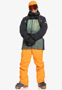 Conjunto de esquí que incluye una chaqueta negra y verde con una cremallera naranja, combinada con pantalones de un naranja brillante. Accesorios: un gorro naranja brillante y gafas de esquí.