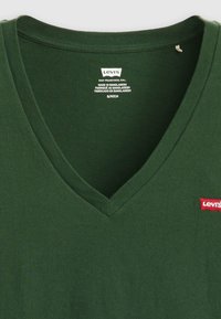 Green v-neck t-shirt made of cotton, featuring a red Levi's logo patch. The fabric has a smooth texture, with a relaxed fit and straight seams.