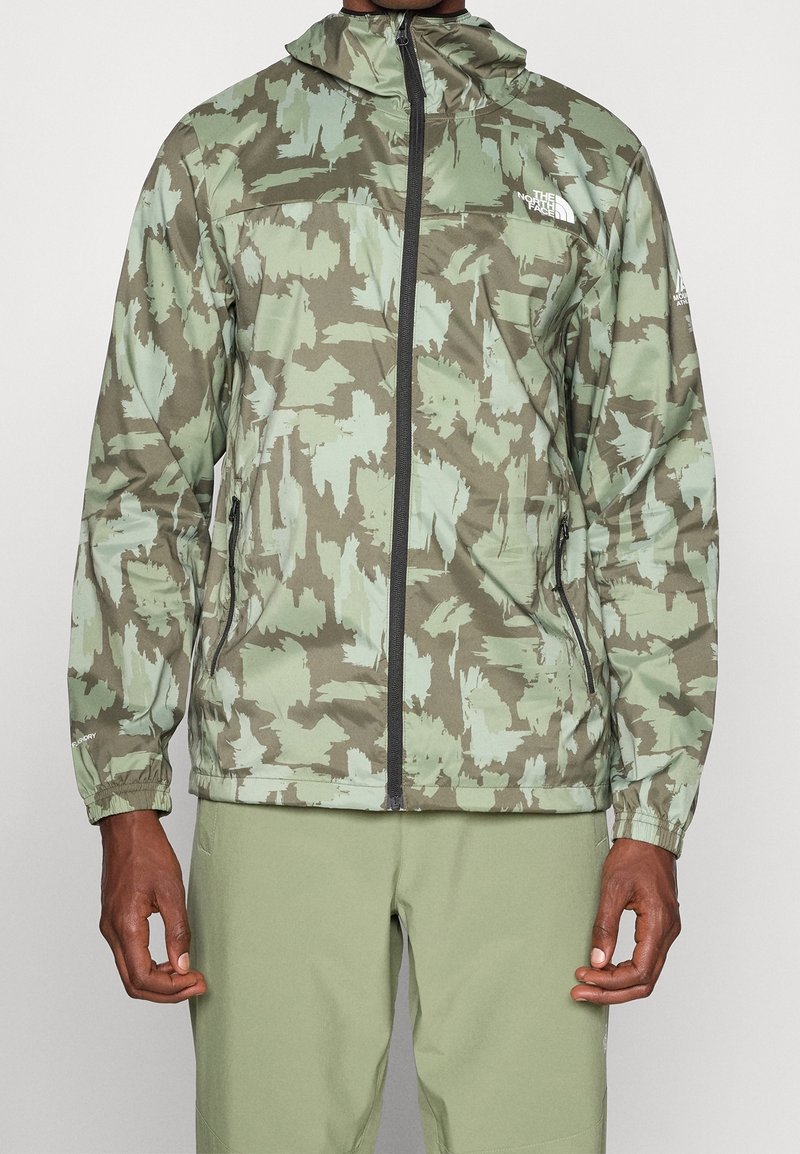 Personne portant une veste à capuche zippée camouflage verte et marron avec des poignets élastiques et des poches avant zippées.