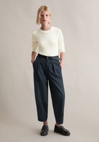 Hellgelber, figurbetonter Pullover mit langen Ärmeln, kombiniert mit dunkelblauen, plissierten Hosen und schwarzen Leder-Loafern, vor neutralem Hintergrund.