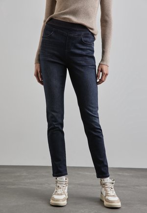 Persoon die donkere skinny jeans draagt, een beige geribde longsleeve top en lichtbeige hoge sneakers, staand op een grijze vloer.