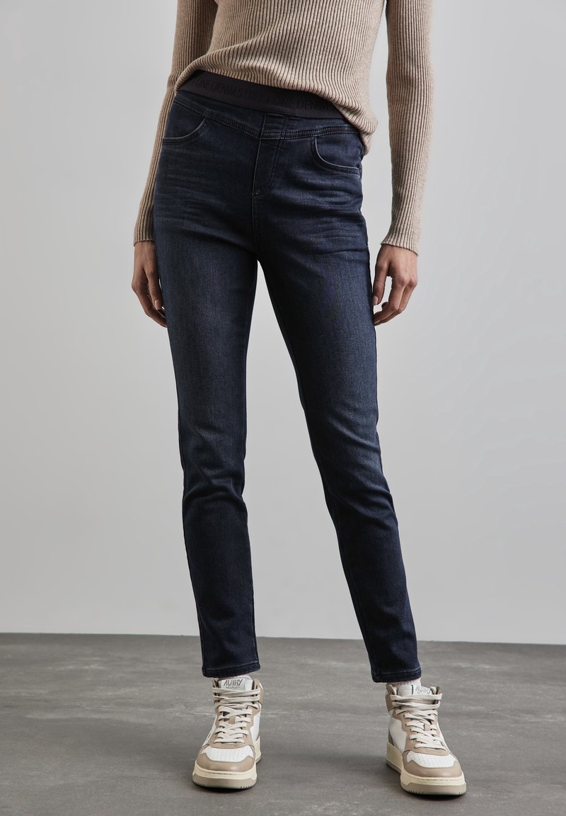 Personne portant un jean skinny foncé, un haut à manches longues côtelé beige et des baskets montantes beige clair debout sur un sol gris.