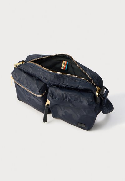 Τσαντάκι ώμου σε ναυτικό μπλε με δύο μπροστινές τσέπες με φερμουάρ σε χρυσή απόχρωση, άνοιγμα με φερμουάρ στο πάνω μέρος και ρυθμιζόμενο κορδόνι σε λευκό φόντο.
