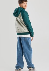 Giacca a blocchi di colore con cappuccio e maniche verdi e corpo grigio chiaro. Indossata sopra una maglietta bianca e jeans blu morbidi. Calzature casual.