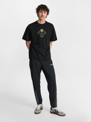 Junger Mann steht mit den Händen hinter dem Rücken, trägt ein schwarzes T-Shirt mit Grafik, schwarze Trainingshose und graue Sneakers vor weißem Hintergrund.