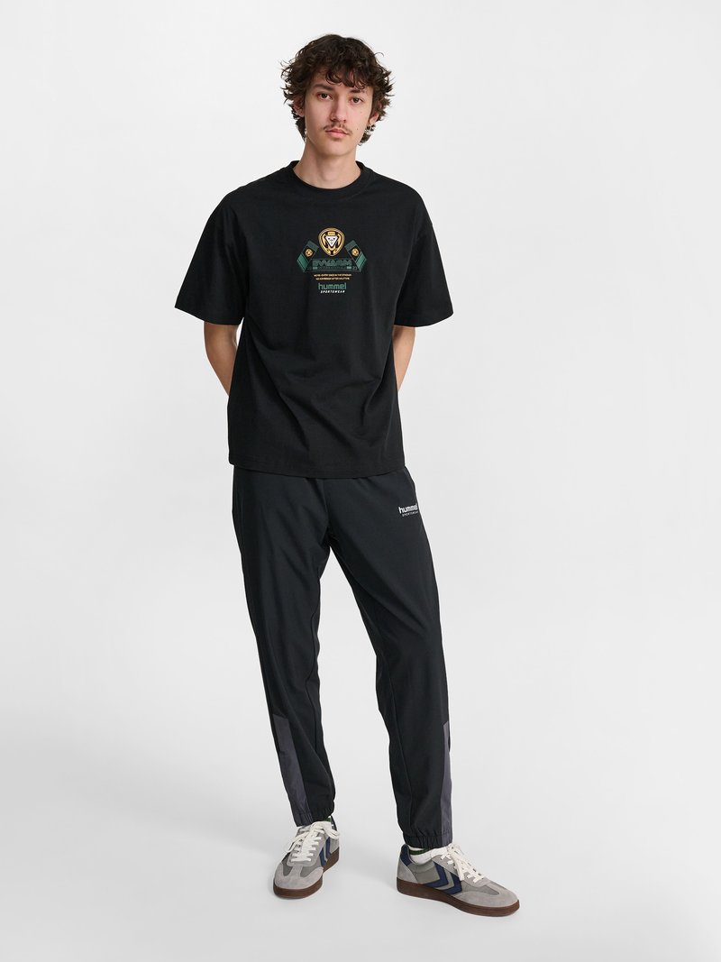 Junger Mann steht mit den Händen hinter dem Rücken, trägt ein schwarzes T-Shirt mit Grafik, schwarze Trainingshose und graue Sneakers vor weißem Hintergrund.