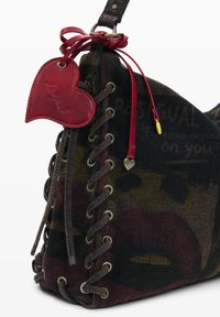 Borsa di tessuto scuro con lati allacciabili, etichetta in pelle a forma di cuore rossa e piccolo charm a forma di cuore in metallo su un cordino rosso.