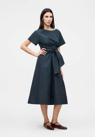 Γυναίκα με μακριά σκούρα μαλλιά που φοράει ένα σκουρομπλε μίντι φόρεμα με κοντά μανίκια και δεμένη μέση, συνδυασμένο με σκούρα κόκκινα μπαλαρίνες.