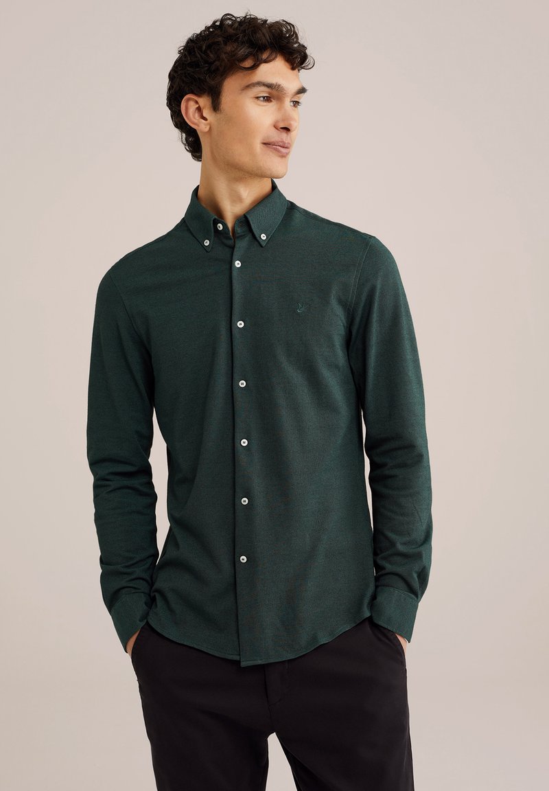 Tumši zaļš krekliņš ar garām piedurknēm, ar apkaklīti, baltām pogām un nelielu logo uz krūtīm, izgatavots no struktūrētas auduma.