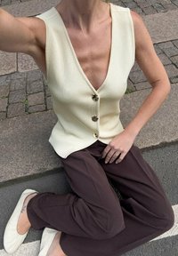 Geribbelde, mouwloze, crème kleurige top met een diepe V-hals en gouden knopen, gecombineerd met loszittende, bruine broeken en lichtgekleurde flats.