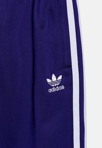 Shorts de sport violets avec des rayures blanches sur les côtés, arborant un logo Adidas brodé en blanc sur la zone de la poche gauche. Texture lisse.