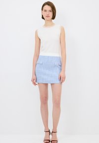 Valkoinen hihaton toppi yhdistettynä vaaleansinihtee huivikankaasta valmistettuun minihelmaan, jossa on kaksi etutaskua ja kultaiset napit. Asusteena ruskeat korot.