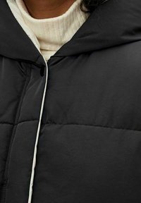 Doudoune noire à capuche, avec un design matelassé. Détail de fermeture à boutons et cordon de serrage blancs visible. Texture douce et mate.