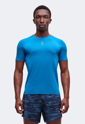 Homme portant un t-shirt de sport ajusté bleu vif à manches courtes et un short bleu foncé à motifs, debout, face à l'avant avec une expression neutre.
