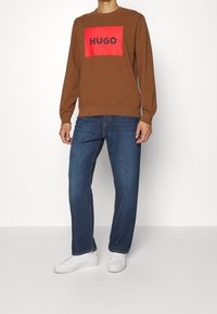 Sweat-shirt marron avec un logo rectangulaire rouge portant l'inscription « HUGO », associé à un jean bleu et des baskets blanches ; coupe décontractée avec poignets côtelés.