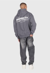 Mann, der einen grauen Hoodie von DROPSIZE CO. und verblasste schwarze Jeans trägt, steht mit dem Rücken zur Kamera und hat eine Hand in der Tasche vor einem schlichten Hintergrund.