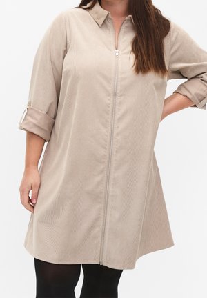 Vrouw draagt een beige lange mouwen jurk met rits en opgerolde mouwen en zwarte legging tegen een effen witte achtergrond.