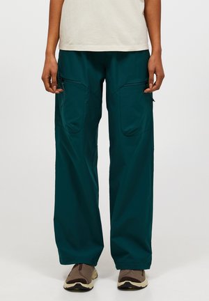 Person iført løse mørkegrønne cargobukser med lynlåslommer på lårene, hvid top og brune vandresko, stående mod hvid baggrund.