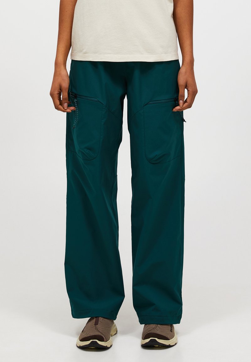 Persona vestida con pantalones cargo verdes oscuros y holgados con bolsillos con cremallera en los muslos, camiseta blanca y zapatos de senderismo marrones, de pie frente a un fondo blanco.