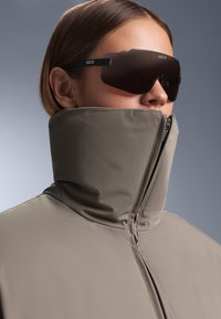 Szürke, magas nyakú kabát cipzárral és sima textúrával, nagy sötét napszemüveggel párosítva, amely merész geometriai mintázattal rendelkezik.