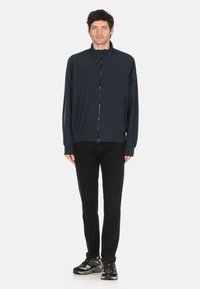Navy blue lightweight jacket with a high collar, zip front, and side pockets, paired with black pants and multi-patterned shoes.