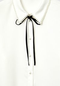 Camicia bianca con colletto decorato da perle, fiocco nero e bottoni perlacei lungo la patta. Tessuto dalla texture liscia.