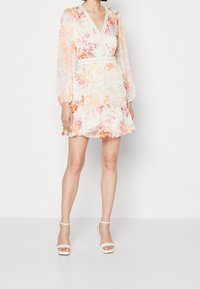 Robe mini fleurie à manches longues, décolleté en V et ourlet à volants. Confectionnée en tissu léger, présentant des nuances de pêche, rose et crème.