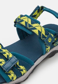 Turkos och gulmönstrad justerbar sandal med tygremmar, vadderad fotbädd och texturerat mönster för grepp. Gummiyttersula, robust design.