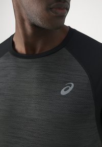 Short-sleeve athletic shirt in dark gray with black accents, featuring a textured finish and a small reflective logo on the left chest.