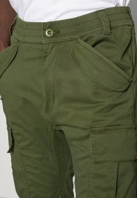 Grüne Cargo-Hose aus strapazierfähigem Gewebe, mit mehreren Taschen, einem taillierten Bund und sichtbaren Nähten. Strukturierte Oberfläche.