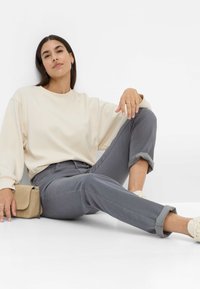 Cremefarbener Langarm-Sweater kombiniert mit hochgerollten grauen Slim-Fit-Hosen. Beigefarbene Umhängetasche mit Kette und hellen Schuhen. Minimalistisches Design.