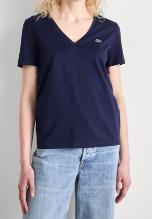 Woman wearing a navy blue V-neck T-shirt with a small green crocodile logo and light blue jeans against a plain background.