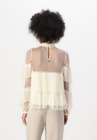 Sheer cream blouse with layered ruffles, decorative stitching, a high neck, and a back keyhole opening. Translucent texture enhances detail.