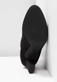 Chaussure à talon haut en daim noir avec un bout pointu et une semelle texturée. Vue arrière montrant un talon lisse et un logo discret gravé sur la semelle.